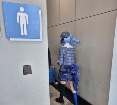 图片中一位蓝发双马尾的coser正背对镜头站在男厕所门口。她穿着灰色格子的水手服校服，戴着同系列的帽子，背后系着一个蓝色大蝴蝶结，搭配黑色过膝袜和蓝色长筒靴。左侧墙上挂着蓝色的男厕所标识，上面有白色的男性符号，下方还有一个小型的男厕所指示牌。背景是灰色的墙面，地面上能看到一个蓝色的垃圾桶和一些散落的物品。