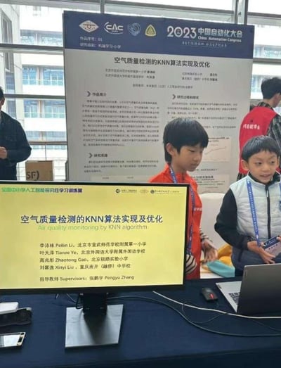 2023中国自动化大会现场，两名中小学生站在展台旁展示科研项目。展台上的电脑屏幕和背景展板均显示项目标题“空气质量检测的KNN算法实现及优化”，屏幕上列出了来自北京、重庆多所中小学的学生作者及指导教师姓名。背景可见大会场馆的玻璃窗和其他参会人员，整体场景体现了青少年在学术会议中展示人工智能与环境监测交叉领域研究成果的画面。