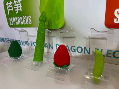 图片展示了一组创意造型的电子科技产品，以蔬菜和水果为设计灵感，包括绿色芦笋造型、红色草莓造型、绿色豌豆荚造型和绿色牛油果造型的物品，均放置在透明亚克力展示架上。背景中有带有“芦笋 ASPARAGUS”字样的绿色标牌，以及部分可见的公司名称“道格电子科技有限公司”和英文“FO...AN DRAGON ELEC...”，推测是该公司在展会或产品发布会上的产品陈列场景。