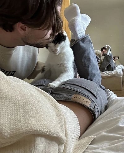 画面中，一名留着胡须、穿着灰色卫衣的男子正侧身躺着，双腿微抬，白色袜子清晰可见。他的腿上坐着一只黑白相间的猫咪，男子低头温柔地亲吻猫咪的头部，猫咪则眼神平静，略带一丝慵懒的冷漠，前爪自然搭在男子的牛仔裤上。男子所穿的灰色牛仔裤后口袋处有一个棕色皮质标签，上面印有“NV-KD”字样。背景中，另一名男子同样双腿朝上仰卧在床铺上，手中似乎抱着一把吉他，营造出舒适惬意的居家氛围。整体场景充满生活气息，展现了人与宠物相处的温馨瞬间以及朋友共处的轻松日常。