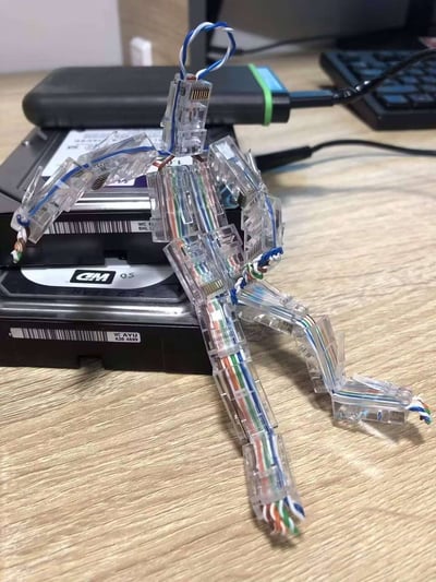 一张展示创意DIY作品的照片。图中是一个用人形结构排列的网线水晶头和彩色网线组成的机器人造型，它被放置在几个叠放的硬盘上。这个手工制品展现了将普通网络配件转化为艺术创作的巧妙构思，水晶头构成了机器人的关节和身体框架，彩色网线则作为肌肉和连接线，整体造型栩栩如生。背景中隐约可见电脑键盘等办公设备，暗示这可能是出自技术人员的创意作品。