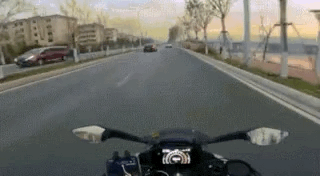 这是一段第一视角拍摄的动态GIF，画面展示了驾驶摩托车在城市的宽阔道路上行驶的场景，道路两侧有居民楼和行道树，前方还有其他行驶的车辆，整体呈现出日常骑行通勤的氛围，画面流畅，能感受到骑行时的速度感。