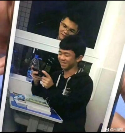 这是一张利用巧合的视觉错位制造搞笑效果的图片：前景是一位穿着黑色连帽衫的男生，正双手拿着手机低头专注操作，脸上带着开心的笑容，看起来像是在上课或自习时摸鱼；而他身后的窗户处，另一位戴眼镜的男生的脸出现在画面中，从拍摄视角看仿佛是从玩手机男生的“头顶”探出来，形成一种“被人悄悄盯着摸鱼”的滑稽既视感，完美还原了上课摸鱼时担心被抓的微妙又搞笑的氛围。