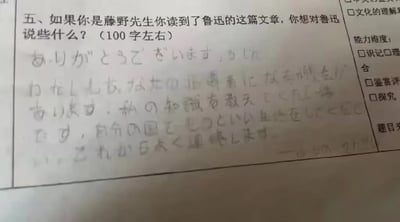 一张语文试卷的局部实拍图，主要展示第五道试题及手写答案。试题内容为“如果你是藤野先生你读到了鲁迅的这篇文章，你想对鲁迅说些什么？（100字左右）”，下方是用日语手写的回答，字迹略显潦草，内容疑似藤野先生对鲁迅表达感谢与期许，提及希望传授的知识能成为鲁迅为国效力的力量，并愿保持联系。