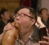 图片中，一位戴着眼镜的中年男子正身处热闹的餐厅环境中，左手紧握一只超大鸡腿，正张大嘴巴用力啃食，表情十分投入陶醉。周围可见其他食客，背景嘈杂充满生活气息，右手似乎还拿着手机，整体画面展现出一种随性享受美食的场景。