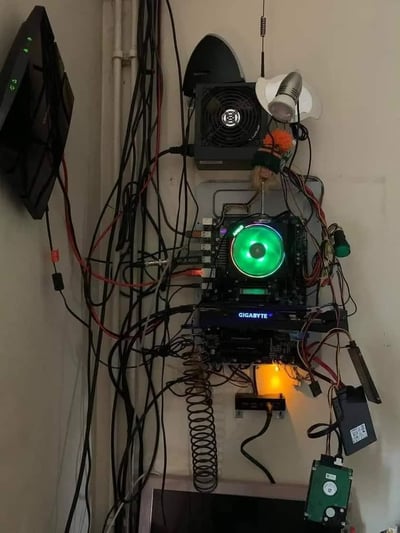 这张图片展示了一个独特的DIY电脑安装方案，将电脑组件直接安装在墙上而非传统机箱中。画面中央是一块主板，配有一个亮绿色的散热风扇，下方可见GIGABYTE品牌标识。各种颜色的线缆（黑色、红色为主）从组件中延伸出来，显得有些杂乱无章。安装在主板上方的是一个黑色散热风扇，旁边还有一个小型天线和其他电子元件。整体呈现出一种极客风格的DIY精神，展示了电脑硬件的原始美感与功能性的结合。
