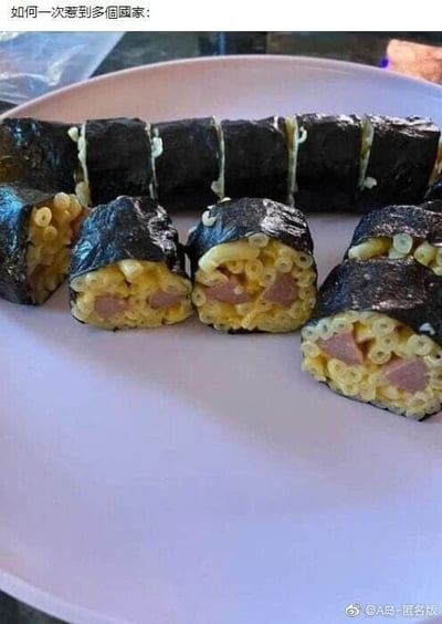 这是一张静态原始拍摄图，展示了一盘混搭风格的黑暗料理：制作者模仿日本寿司卷的外观，用海苔作为外皮，内部却填充了美式餐食中常见的通心粉和火腿，完全替换了寿司传统的米饭、刺身、腌菜等食材。图片顶部配有文字“如何一次惹到多个国家：”，这个调侃的核心在于，这种不伦不类的混搭既违背了日本寿司的传统制作逻辑，又将通心粉改造成了不符合欧美常规食用场景的形态，以此玩笑式地吐槽该食物会同时冒犯到喜爱传统寿司的日本民众，以及习惯常规通心粉吃法的欧美国家民众，充满了搞笑的吐槽趣味。