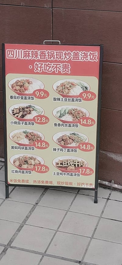 这是一张户外实拍的立式餐饮价目展示牌照片，展示牌为黑框立式结构，顶部红色背景区域用白色字体标注店铺主营品类“四川麻辣香锅现炒盖浇饭·好吃不贵”；下方米黄色底色区域分左右两列，依次展示8款盖浇饭的菜品示意图、名称及定价，价格区间从9.9元到17.8元，属于平价大众餐品；展示牌最底部用黑色字体标注福利信息：“米饭免费续，热汤免费喝，现炒现做·好吃不贵”。展示牌放置在浅灰色方格地砖上，后方背景为棕色波纹墙面。