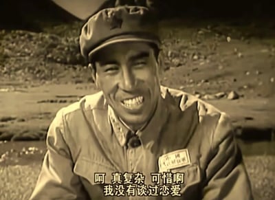 一张黑白老照片风格的影视截图，画面中是一位穿着军装的男性，戴着军帽，面带笑容。他坐在户外的自然环境前，背景有山石和植被。人物胸前佩戴着写有名字的标牌。图片下方有字幕："呵 真复杂 可惜啊 我没有谈过恋爱"。