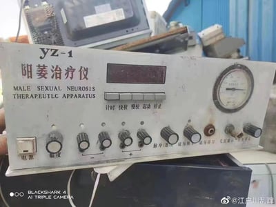 图片展示了一台陈旧的医疗设备，型号为YZ-1的阳痿治疗仪。设备主体为白色，表面有明显的污渍和使用痕迹，显示其年代久远。仪器正面有中文和英文标识，中文名称为"阳痿治疗仪"，英文翻译为"MALE SEXUAL NEUROSIS THERAPEUTLC APPARATUS"（注意英文存在拼写错误，应为THERAPEUTIC）。设备配备了数字显示屏、压力表、多个旋钮和按钮，以及各种接口。按钮上标有"计时、快校、慢校、起动、停止"等功能，旋钮标注了"电源、幅度、脉冲/连续、磁场频率"等调节选项。设备被放置在一堆废弃物品中，背景中可以看到其他旧电器和蓝色的金属棚屋，整体呈现出一种被遗弃的状态。