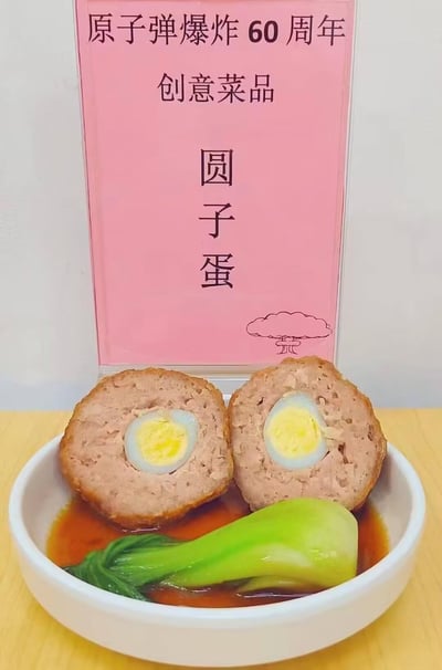 这是一道为纪念原子弹爆炸60周年设计的创意菜品「圆子蛋」：白色碗中摆放着被切成两半的肉圆包裹水煮蛋的料理，搭配青菜与酱汁，后方立着粉色标牌，标注了主题、菜品名称，还配有原子弹爆炸蘑菇云的简笔画。
谐音梗说明：「圆子蛋」谐音「原子弹」，将肉圆包蛋的家常料理与纪念主题结合，是充满脑洞的玩梗创意菜。