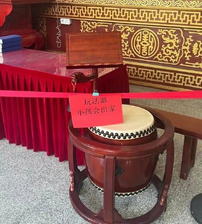 图片展示了一个具有传统中式装饰风格的室内场景，背景是带有金色龙纹和“囍”字图案的红色墙面。前景中有一个木制鼓架，鼓身呈棕红色，鼓面为白色并带有黑色装饰。鼓的上方用红色纸牌悬挂着一条警示标语，上面写着“玩法器 小孩会出家”。鼓的周围用红色绸带围起，形成隔离区域。整体场景给人一种传统寺庙或中式礼堂的氛围，标语内容带有民间幽默和调侃的意味，暗示不要让小孩随意触碰宗教法器。