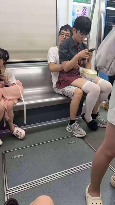 这是在地铁车厢内的真实场景照片，画面右侧的白T恤男士与穿女装（黑衬衫、红格裙、白长筒袜）的路人相邻而坐，因视觉错位，看起来像是男士从背后环抱住女装路人，制造出亲密的假象，实际两人是分开的。画面左侧还有一位扎双马尾的女生在低头看手机，整个场景因视觉错位显得十分迷惑搞笑。