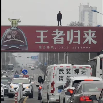 这是一张现实拍摄的城市道路场景照片，画面上部是横跨道路的红色大型商业广告牌，广告牌左侧印有百年兰陵王白酒的产品图案，右侧配有硕大的白色文字“王者归来”，下方标注服务热线0539 5588998。一名身穿黑色衣物的男子正站在广告牌的顶部边缘，手中拿着白色物品。广告牌下方的城市道路处于拥堵状态，可见多辆社会车辆，还有尾部印有“武装押运”字样的押运车，以及亮着警灯的警车停在道路旁。广告牌上的“王者归来”文字与站在高处的男子、下方的警务相关车辆形成了极具戏剧感的巧合效果，看起来仿佛文字恰好对应了当下的场景，充满趣味性。