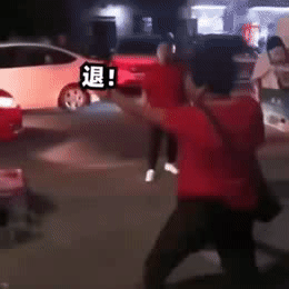 夜间街道场景中，一名身穿红色上衣的男子正做出夸张的后退手势，双臂向两侧张开，身体向后倾斜，同时画面中多次出现“退!”字，配合其动作增强表达效果。背景可见停放的汽车和模糊的行人，地面散落着一些物品，整体氛围带有戏剧性和搞笑感，仿佛在驱赶或拒绝某物。