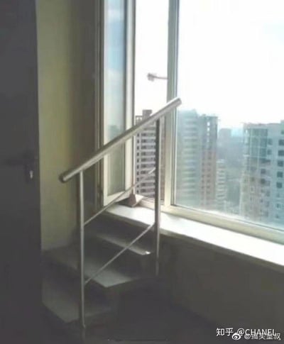 这张图片展示了一处室内的奇葩装修设计：在靠近窗户的位置安装了一段带有金属扶手的楼梯，然而楼梯的顶端直接对着封闭的窗户，没有任何可通行的出口或平台，完全不具备实用价值，属于典型的反人类迷惑设计，让人不禁吐槽设计师的不合理构思。窗外可以看到城市中的高层住宅楼，显示这是一处高层建筑内的空间。