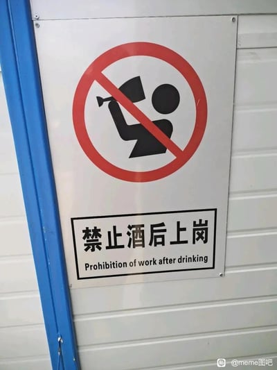 这是一张拍摄于板房墙面的安全警示标识牌照片，标识牌为金属质感的白色底板，上方是国际通用的红色圆圈加斜杠的禁止标志，内部绘制了一个举着类似酒杯物品的黑色卡通人形，明确传达禁止的行为；下方是黑框包裹的文字区域，上方是加粗的中文“禁止酒后上岗”，下方是对应的英文翻译“Prohibition of work after drinking”。该标识用于在工厂、工地等作业场景中，警示工作人员严禁饮酒后上岗作业，避免因酒精影响操作引发安全事故。