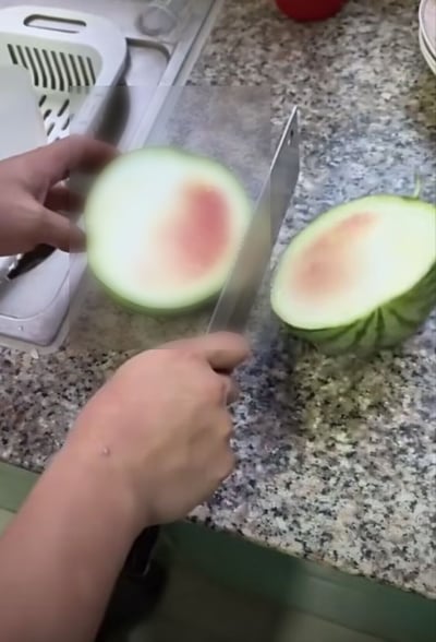 这张实拍图呈现了厨房内的场景，花岗岩纹理的台面上放着被切成两半的西瓜，该西瓜仅有中心非常小的一块区域是淡红色的成熟瓜瓤，其余绝大部分面积都是青白色的未成熟组织，一只手正握着菜刀对准其中一半西瓜，另一只手扶着西瓜，背景可见水槽旁的白色滤水篮，整体展现出买到未成熟西瓜的意外、哭笑不得的场景。