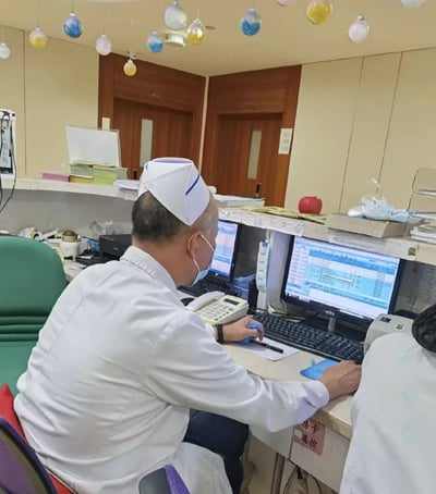 医院办公室场景中，一位身着白色大褂、头戴护士帽并佩戴口罩的医护人员正坐在办公桌前专注工作。桌上摆放着多台电脑显示器，显示着医疗相关数据表格，其右手持笔在纸上记录，左手操作键盘。办公桌周围有电话机、文件、绿色座椅等办公物品，背景可见木质房门（标有“205”字样）及悬挂的彩色球形装饰，整体呈现出医院办公区域的日常工作氛围。