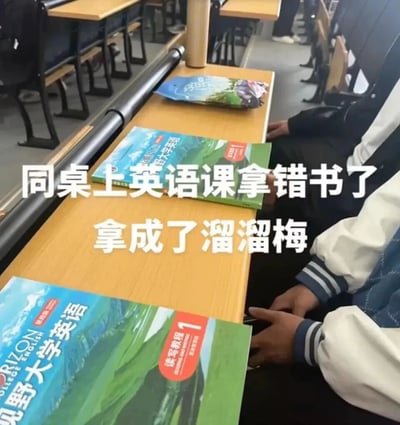这是一张在大学教室内拍摄的真实场景图片，画面展示了多排课桌和坐在桌前的学生。其中一名学生在上英语课时出现了乌龙：他误将溜溜梅的食品包装当成了《视野大学英语·读写教程1》课本带到了课堂上，两者的封面配色、设计风格高度相似，才导致了这个有趣的错误。图片上的白色文字标注点明了这个笑点，让整个场景充满了校园生活的轻松搞笑氛围，也因为包装和课本的高度相似性容易引发大学生的共鸣。