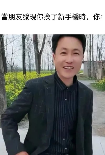 这是一张网友创作的趣味梗图，顶部配有繁体中文文案“當朋友發現你換了新手機時，你：”，下方是一位身处乡间户外场景（背景有油菜花田、乡间小路）的男士，他穿着深色西装外套搭配条纹衬衫，脸上露出极其灿烂、喜悦的笑容，用这种夸张的开心表情来对应换了新手机被朋友发现时，自己内心那种藏不住的小得意与快乐，将日常小事里的情绪放大，容易引发网友的共鸣，适合用来调侃生活里的小确幸。