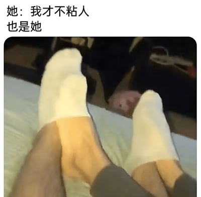 图片上方配有文字“她：我才不粘人 也是她”，下方是一双穿着白色袜子的脚放在床上的特写镜头，床底下有一只小型宠物（可能是仓鼠或豚鼠）探出头来张望。通过文字与画面的结合，幽默地表现了人们嘴上说不粘人但实际行为却很粘人的反差状态。