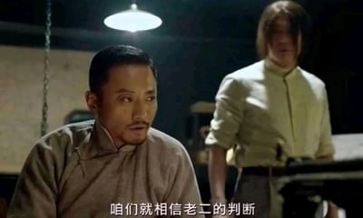 这是一张出自电影《让子弹飞》的静态截图，画面呈现了一个昏暗的室内议事场景：上方悬挂的一盏亮着的吊灯是主要光源，前景处一位穿着灰色中式立领长衫、发型整齐的男子坐着，面露思索神情，嘴巴微张，正在发言；他身后站着一位留着中长发、穿着浅色上衣的男子，身影处于稍暗区域，姿态沉稳。画面底部配有白色字幕“咱们就相信老二的判断”，传递出剧中角色们商议行动方案时，决定信任弟兄“老二”的判断的情节，整体氛围严肃凝重，充满了帮派议事的紧张感。