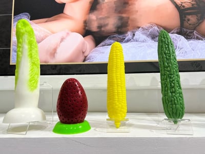 图片展示了一个创意艺术装置，前景中有四个造型独特的蔬菜形状物体，从左到右分别是生菜造型、草莓造型、玉米造型和苦瓜造型，每个物体都被放置在透明支架上。背景是一幅模糊处理的人物图像，形成了鲜明的视觉对比。这种将日常蔬菜与人体艺术并置的方式，可能意在探讨自然与人体、食物与欲望等主题，具有一定的讽刺和幽默效果。