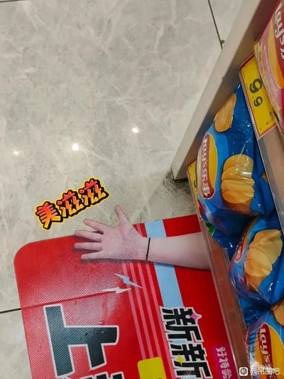 这是一张在超市内拍摄的趣味视觉错位照片，画面右侧的货架上摆放着标价9.9元的蓝色包装乐事薯片，货架旁的红色地垫上印有一只手的图案，恰好与旁边露出的人的手臂完美重合，营造出“手从地垫里伸出来”的错觉效果，画面中还添加了“美滋滋”的黄色趣味贴纸，让整幅图充满搞笑氛围。