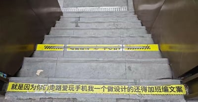 这张图片展示了一段地铁或商场的楼梯，在台阶下方贴有醒目的黄色警示标语。标语用戏谑的语气写道：“就是因为你们走路爱玩手机我一个做设计的还得加班编文案”，幽默地吐槽现代人走路玩手机，设计师不得不加班想出这些警示语。整体风格诙谐幽默，带有些许无奈和自嘲。

此语句包含自我调侃和对常见社会现象的吐槽，有一定的双关和玩梗，既提醒行人注意安全，也顺带表达了设计行业的辛苦。