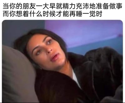 这是一张适配日常社交场景的表情包，属于文字加注的影视截图类内容。图上方配有文字“当你的朋友一大早就精力充沛地准备做事而你想着什么时候才能再睡一觉时”，图中是真人秀明星金·卡戴珊，她躺在床上，裹着深色毛绒外套，眼神涣散、表情倦怠，整个人呈现出强烈的困意和慵懒感，精准地还原了很多人在朋友充满干劲早起做事时，自己却只想补觉、提不起精神的反差状态，非常贴合休息日或早起时的真实心态，容易引发共鸣。