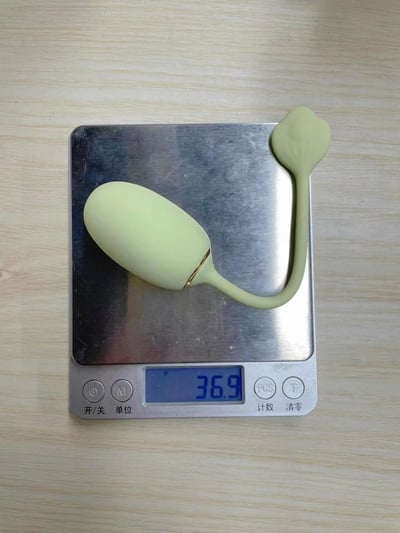 一张静态照片，展示了一个浅绿色的振动器放在电子秤上，电子秤显示重量为36.9。电子秤放置在浅色木质桌面上，秤的表面有使用痕迹。振动器设计成椭圆形主体带一个弯曲的底座。电子秤上有四个按钮，分别标注着'开/关'、'单位'、'计数'和'清零'。