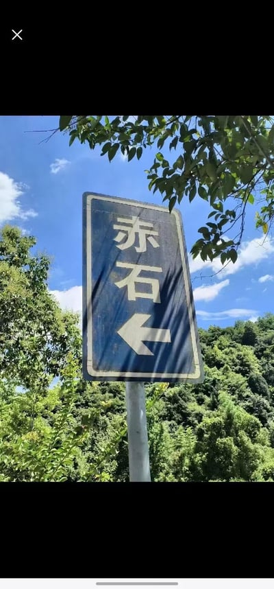 一张户外实景拍摄的路标照片。画面中央是一块蓝色路牌，边缘有白色边框，牌面上方用白色简体中文写着'赤石'二字，下方是一个向左的白色箭头，指示前往赤石的方向。路牌后方是茂密的绿色树木，枝叶部分遮挡了天空，背景可见晴朗的蓝天和几朵散落的白云，阳光透过树叶在路牌上形成斑驳的光影效果，整体氛围清新自然。
