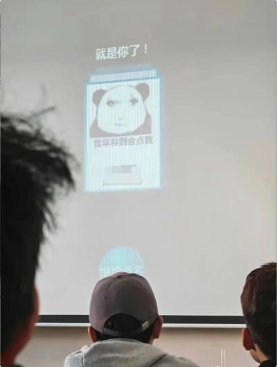 这是一张记录课堂或职场培训场景的静态照片，前景是三位背对镜头的参与者，其中中间的人戴着灰色棒球帽，他们正共同注视前方的投影屏幕。屏幕上展示的是一个趣味点名系统界面：顶部配有文字“就是你了！”，核心区域是一个经典的熊猫头表情包，表情包下方配文“我早料到会点我”，表情包下方有一处被打码的区域，界面底部还有“继续点名”的按钮。这个界面将严肃的点名环节和搞笑的网络表情包结合，让原本枯燥的考勤流程变得诙谐有趣，缓解了参与者的紧张感，也体现了职场或校园中轻松的文化氛围。