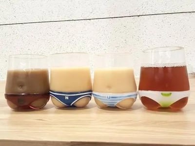图片展示了四个透明玻璃杯整齐排列在木质桌面上，背景为白色纹理墙面。每个玻璃杯中部都有模拟内裤的造型设计：从左到右依次为棕色饮品配纯色系内裤造型、米色饮品配蓝色带“N”字内裤造型、米色饮品配蓝白条纹内裤造型、红褐色饮品配绿色装饰内裤造型。整体设计将日常饮具与幽默元素结合，视觉上形成趣味反差。