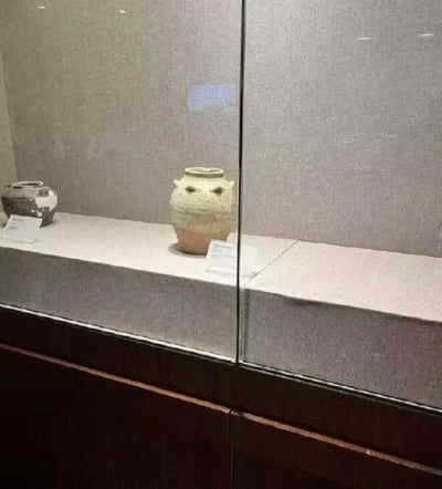 这是博物馆展柜中的场景，展台上摆放着两件古代陶罐，中间的陶罐因展柜玻璃的反光，搭配自身的双系耳，恰好形成了类似眼睛和嘴巴的视觉效果，看起来像一张带着怒气的脸，让原本古朴的文物意外拥有了搞笑的萌感，形成了趣味的反差感。
