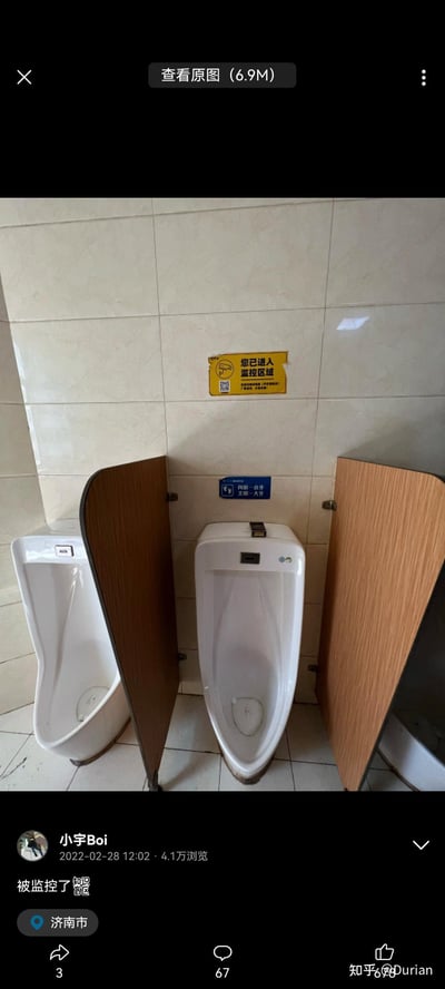 这是一张来自知乎的社交媒体截图，展示了济南某场所的男卫生间场景：墙面上铺着米白色瓷砖，两个白色小便池之间设有木质隔板，在小便池正上方的墙面上，贴着一张黄色的“您已进入监控区域”警示标识，下方还有蓝色的“向前一小步 文明一大步”文明标语。在私密的如厕区域设置监控标识，打破了人们对公共场所隐私的认知，会让人感到尴尬和不适，引发对公共设施隐私边界的讨论。