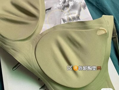 图片为浅绿色内衣的近距离展示，聚焦于内衣的布料纹理和设计细节，突出其“可拆卸胸垫”的特点，配有✅和🐻表情符号增强视觉提示。背景中隐约可见带有线条图示的纸张，可能为产品说明或包装的一部分，整体构图清晰，光线柔和，展现了内衣的材质质感。