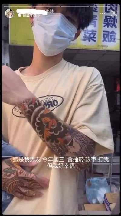 这是一张来自中国台湾地区社交媒体的视频截图，画面主体是一位戴着白色口罩、穿着米色T恤的男生，他的手臂和手部有大面积风格偏向社会青年的纹身。画面下方配有一段充满强烈矛盾感的文字：“这是我男友，今年初三，会抽烟、改车、打我，但我好幸福”。这段文字将“初三学生”“抽烟”“改车”“家暴”这些负面标签与“幸福”的感受结合，制造出荒诞的反差效果，或是用来讽刺扭曲的恋爱观，或是网友为制造话题编造的段子，以此在网络上引发讨论。背景是带有繁体中文的街头店铺招牌，进一步佐证了场景位于台湾地区。