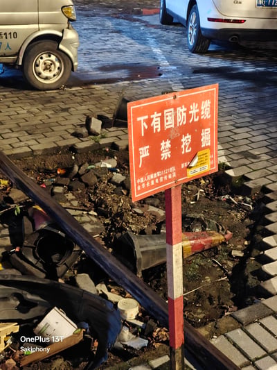 这是一张夜晚拍摄的户外实拍照片，画面前景是一块竖立在红白相间支撑杆上的橙色警示牌，上面明确标注“下有国防光缆 严禁挖掘”，警示牌后方是已经被开挖的地面，散落着碎石块、泥土、破损的管线部件和垃圾，周边是铺设好的砖面人行道，路边还停放着两辆汽车。场景呈现出施工进行到一半突然停工的状态，刚好贴合标题“怎么挖一半不挖了”的疑问，由于损毁国防光缆后果十分严重，施工方大概率是看到警示牌后立刻停止了作业。