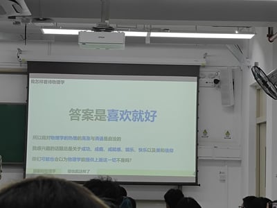 这是一张高校课堂的实拍图，画面核心是投影幕布上的PPT内容，主题为「我怎样看待物理学」，核心观点是「答案是喜欢就好」。内容中提到分享者对物理学的热情高涨与消退是自洽的，自身感兴趣的是成功、成瘾、成就感、娱乐、快乐以及美和信仰这类话题，调侃听众可能会以为物理学能提供这些，最后用「眼前的物理学，但也就这样了」收尾，带着佛系又略带调侃的态度分享对物理学的看法。画面中还能看到天花板上的爱普生投影仪、教室风扇，以及下方听课学生的头顶，还原了真实的课堂场景。