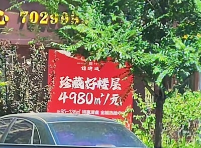 图片展示了一处房产广告场景。画面中央是一个红色背景的大型广告牌，上面用白色大字写着“珍藏好楼层 4980元/㎡”，下方还有“95-136㎡特惠清盘 全城速抢中”的小字。广告牌后方有一栋建筑物，其外墙上可见部分黄色数字“7029”的标识。广告牌前方停放着一辆深色轿车，周围有茂密的绿色树叶和灌木丛，整体环境显得较为生活化。
