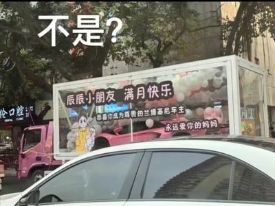 图片展示了一辆装饰有气球的粉色卡车，车身上印有“辰辰小朋友 满月快乐 恭喜你成为尊贵的兰博基尼车主 永远爱你的妈妈”的标语，顶部叠加了醒目的“不是?”文字。背景可见街道树木和“伦口腔”诊所招牌，整体呈现出一种对满月婴儿获赠豪车这一夸张祝福的幽默调侃。