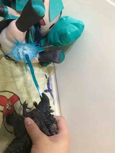 这是一张通过视觉错位与P图特效结合制作的恶搞玩梗图。画面中一只手握着哥斯拉的手办模型，将其头部对准初音未来的毛绒玩偶，同时添加了一道蓝色的“原子吐息”特效，让特效的起点衔接哥斯拉嘴部，终点落在初音未来玩偶的身体上，营造出哥斯拉正用标志性的原子吐息攻击初音未来玩偶的搞笑场景，把两个知名ACG IP结合起来，充满了爱好者的创意恶搞趣味。