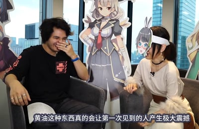 这是一段访谈的视频截图，场景位于室内靠窗的位置，背景是落地窗，窗外可见城市高楼建筑。画面中一男一女坐在灰色沙发上，男士穿着印有红色“FAKKU”字样的黑色T恤，他笑着用手捂住嘴，看起来是在聊到有趣的话题时的反应；女士戴着一个二次元兔耳少女形象的头套，坐在男士右侧。背景中还立着三个二次元美少女风格的人形立牌，风格偏向女仆、兽耳类的二次元设定。画面底部有白色字幕：“黄油这种东西真的会让第一次见到的人产生极大震撼”，这里的“黄油”是谐音梗，指代美少女成人向游戏（“黄游”的谐音，也对应英文“eroge”的谐音）。