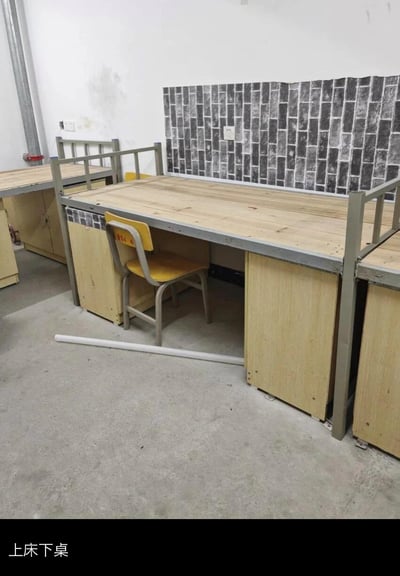 这是一张实拍的集体宿舍场景图，画面中展示了多组金属框架搭配木质桌面的上床下桌连体家具，书桌下方配有储物柜，搭配黄灰配色的简约靠背椅。地面为水泥地，墙面部分区域贴有仿砖纹装饰贴纸，地面还放置着一根白色管材，整体配置朴素实用，符合员工宿舍或学生宿舍的典型环境。