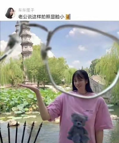一位女子在中式园林背景前，手持眼镜框在镜头前形成圆形框架自拍，利用视觉效果让脸看起来更小。背景中有古塔、池塘荷叶和垂柳，营造出古典园林氛围。