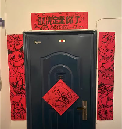 一张展示家庭入户门新年装饰的照片。深蓝色的门上贴有红色菱形“福”字贴纸，福字周围有宝可梦卡通图案。门的正上方悬挂着红色横幅，上面写有“就决定是你了”和“POKÉMON”字样，周围点缀着精灵球、皮卡丘等宝可梦元素。门的左右两侧各有一幅红色竖版春联，左侧春联包含伊布、喷火龙等宝可梦角色，右侧春联则有皮卡丘、卡比兽等形象，整体将传统新年装饰与宝可梦动漫元素结合，营造出喜庆又充满童趣的氛围。