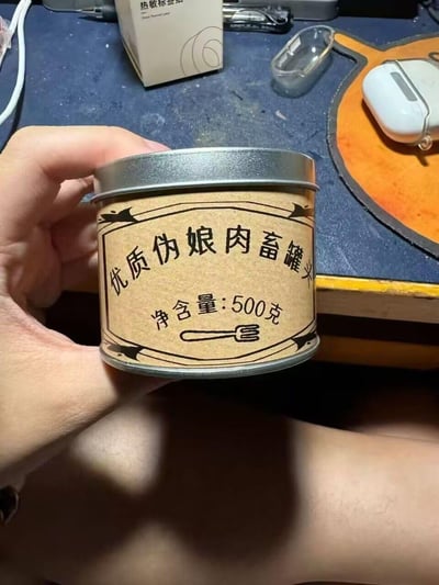 一只手拿着一个金属材质的罐头，罐头表面贴着牛皮纸风格的自制标签，标签上写着猎奇的「优质伪娘肉畜罐头」字样，下方标注净含量500克，还画有一个叉子的简笔画。这是一款利用猎奇名称制造笑点的整蛊道具，不存在实际的该类食品，属于二次元圈子的玩梗创意物品，通过恶搞标签改造普通罐头来达到整蛊、趣味展示的效果。