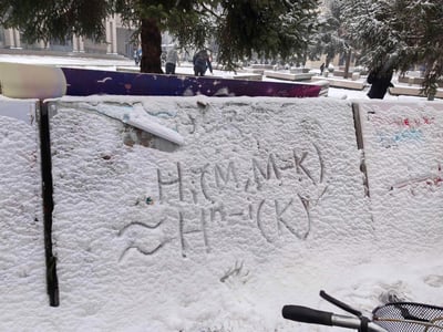 这是一张冬日飘雪的户外实拍图，画面核心是被厚实积雪覆盖的公告板，有人在松软的雪层上手写了「HM-K」「HK」字样，还有一条波浪线图案，公告板上方还有模糊的手写字母。背景里，飘雪的广场/校园区域中，行人在积雪的地面上行走，周围的树木、长椅和建筑都覆盖着白雪，整体氛围充满冬日的清冷与日常的小趣味，没有双关或谐音梗内容。